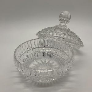 May include: A clear, cut-glass candy dish with a lid. The dish is round with a starburst pattern on the bottom and a diamond-cut design. The lid has a knob and a matching diamond-cut pattern. The dish is open, with the lid slightly ajar.