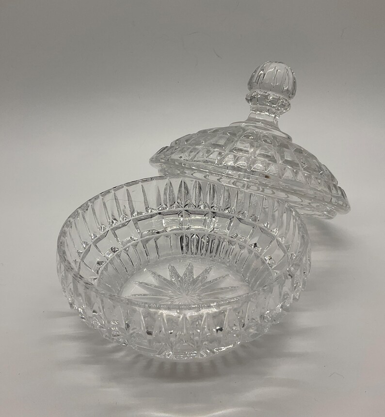 May include: A clear, cut-glass candy dish with a lid. The dish is round with a starburst pattern on the bottom and a diamond-cut design. The lid has a knob and a matching diamond-cut pattern. The dish is open, with the lid slightly ajar.