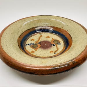 May include: A ceramic bowl with a white glaze and brown, orange, and blue painted designs. The bowl has a wide rim and a deep well.
