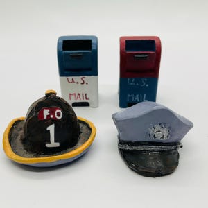 May include: Miniature figurines of a firefighter's helmet, a police officer's hat, and two U.S. mailboxes. The helmet is brown with a yellow brim and the letters "F.O. 1". The mailboxes are blue and red with "U.S. MAIL" text.