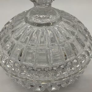 May include: A clear, faceted glass candy dish with a lid. The dish has a round shape with a textured, geometric pattern. The lid features a decorative finial. The glass is transparent, allowing light to pass through.