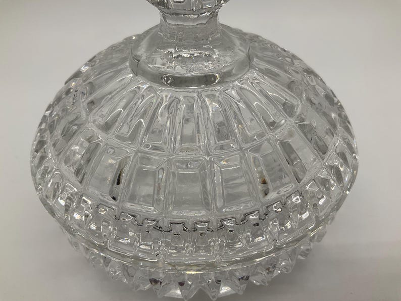 May include: A clear, faceted glass candy dish with a lid. The dish has a round shape with a textured, geometric pattern. The lid features a decorative finial. The glass is transparent, allowing light to pass through.