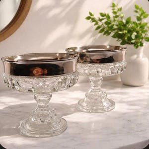 May include: Two clear glass champagne coupe glasses with silver rims. The glasses have a decorative, cut-glass design on the bowl and stem. A white vase with green foliage is in the background.