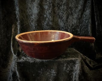 May include: A deep burgundy ceramic bowl with a handle. The bowl has a light brown rim and a white base. The handle is the same color as the bowl. The bowl is likely used for serving or cooking.