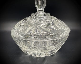 May include: A clear, vintage glass candy dish with a lid. The dish features a starburst pattern and a decorative handle on the lid. The glass is thick and has a reflective quality, suitable for display or serving.