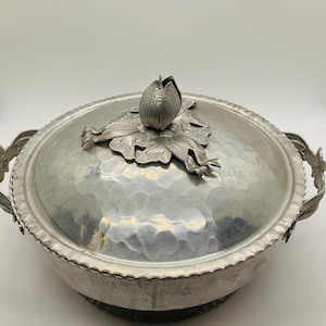 May include: A silver-colored, hammered metal serving dish with a lid. The lid is topped with a decorative flower and leaf design. The dish has handles with a floral design and a scalloped edge.
