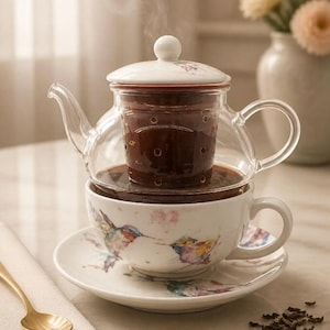 May include: A clear glass teapot with a lid sits atop a white ceramic teacup and saucer. The cup and saucer feature a colorful hummingbird and floral pattern. A gold-toned spoon rests on a napkin nearby, with loose tea leaves scattered on the surface.