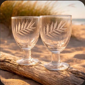 May include: Two clear glass goblets with etched fern leaf designs. The goblets are on a piece of driftwood, with a sandy beach and ocean in the background. The goblets have a classic stem and foot.