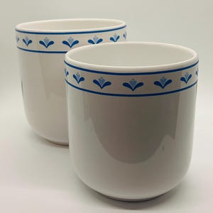 May include: Two white ceramic mugs with a blue floral design around the rim. The mugs have a rounded shape and a glossy finish. The blue floral pattern is a repeating design of stylized flowers.
