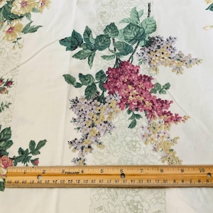 May include: White fabric with a floral pattern featuring pink, yellow, and purple flowers with green leaves. A wooden ruler is placed across the bottom of the fabric, measuring up to 10 inches.
