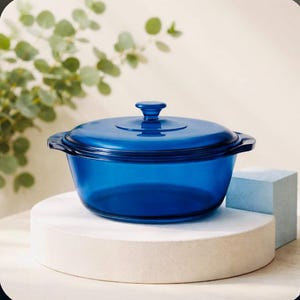 May include: A cobalt blue glass casserole dish with a matching lid. The dish is round with two handles and sits on a white circular platform. The lid has a small knob. The background includes greenery and a light blue cube.