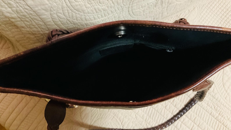 Może przedstawiać: Brązowa sk&oacute;rzana torba typu tote z ciemnym wnętrzem. Torba ma pleciony uchwyt i magnetyczne zapięcie na zatrzask. Wnętrze posiada małą kieszeń. Torba jest ustawiona na neutralnym tle.