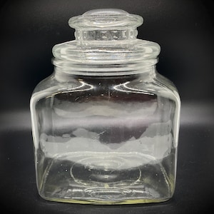 May include: A clear, square-shaped glass jar with a matching lid. The jar has a wide opening and a rounded top, perfect for storing various items. The lid has a decorative, textured design. The jar is empty and sits against a black background.