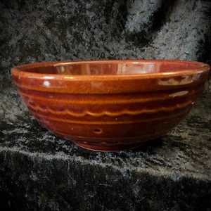 May include: A round, reddish-brown ceramic bowl with a glossy finish. The bowl features horizontal ridges and a decorative scalloped edge. The bowl is likely used for serving food.