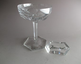 Op de afbeelding: Een heldere kristallen champagnecoupe met geëtste details en een lange steel. Een klein, helder, zeshoekig kristallen stuk met het woord "Baccarat" in zwart schrift staat naast het glas.
