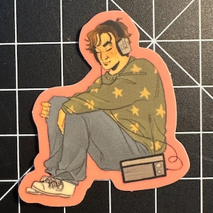 May include: A sticker featuring a person wearing headphones, sitting with their knees up, and listening to a radio. The person is wearing a green sweater with yellow stars and gray pants. The sticker has a pink border.