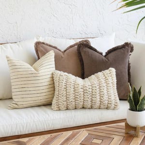 May include: A collection of four decorative throw pillows including one cream pillow with thin horizontal black stripes, one light brown pillow, one dark brown pillow with fringed edges, and one rectangular cream pillow with a chunky knit texture.