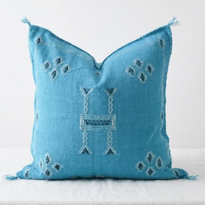 May include: A square, turquoise pillow with a textured weave and decorative stitching. The pillow features geometric patterns in white and dark blue, with fringe details at each corner. The pillow is placed on a white surface.