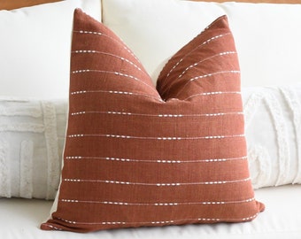 Puede incluir: Un sofá blanco con dos almohadas blancas y una almohada de color óxido con costuras blancas. El sofá está frente a una pared blanca. Una mesa de café de madera con un patrón geométrico está frente al sofá. Una almohada blanca y óxido está sobre la mesa de café.