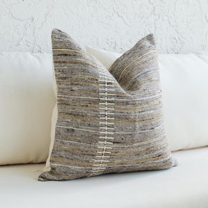 May include: A decorative pillow with a woven textured cover in shades of gray, brown, and cream. The pillow features a vertical white stitched design down the center. It is placed on a white cushion with two other white pillows.