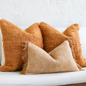 May include: Four decorative pillows in shades of orange and white. Two large, textured orange pillows are behind a smaller, fringed pillow and a white pillow. The pillows are arranged on a white surface.