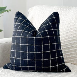 May include: A navy blue decorative pillow with a white grid pattern. The pillow is square and rests on a white textured sofa. The pillow adds a touch of modern style to any living space.