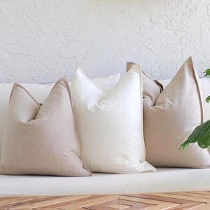 May include: A collection of decorative throw pillows in various shades of beige and white. The pillows have a square shape with a pointed top and a contrasting trim. They are arranged on a white surface, with a wooden element in the foreground.