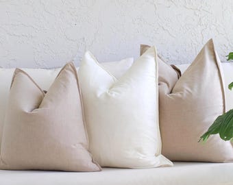 May include: A collection of decorative throw pillows in various shades of beige and white. The pillows have a square shape with a pointed top and a contrasting trim. They are arranged on a white surface, with a wooden element in the foreground.