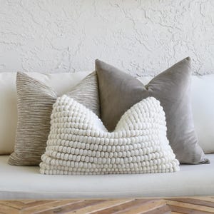 May include: A collection of decorative throw pillows in neutral tones. The arrangement includes a textured white pillow with a bobble design, flanked by a gray and a patterned pillow, and a solid white pillow. The pillows are arranged on a white sofa.