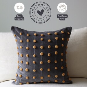May include: A dark grey velvet cushion featuring vertical lines of small, tan pom-poms. The square cushion has a soft, textured look. The words "Handmade with Love" are in a circle above the cushion.