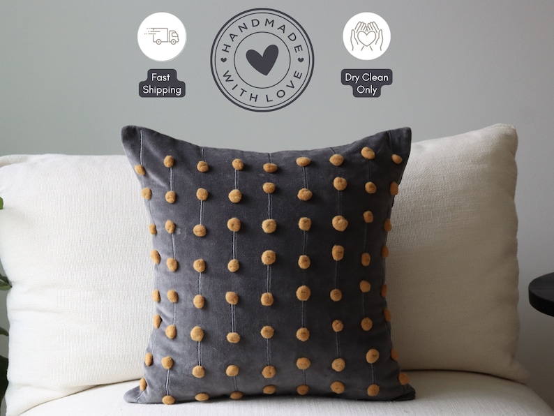 May include: A dark grey velvet cushion featuring vertical lines of small, tan pom-poms. The square cushion has a soft, textured look. The words "Handmade with Love" are in a circle above the cushion.