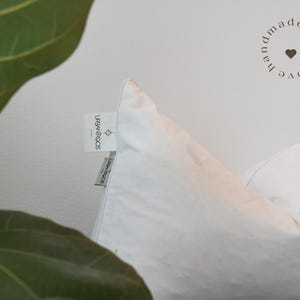 May include: Close-up of a white pillow with a tag that reads "URBAN ROOTS". The pillow is against a white background. The words "handmade with love" are in a circle on the upper right.