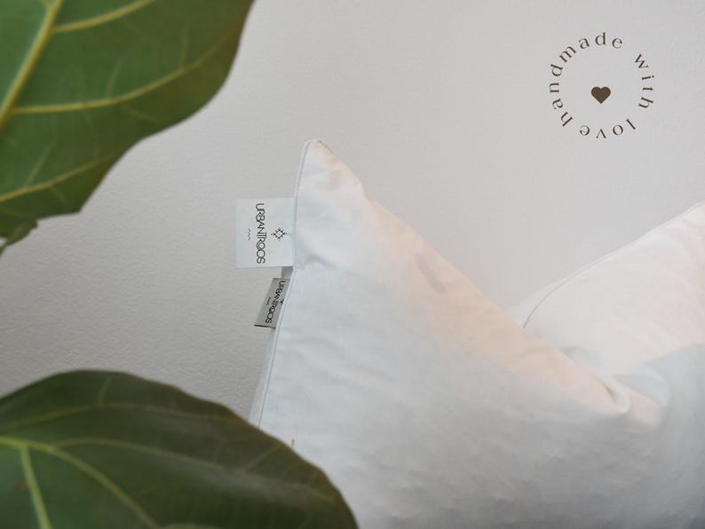 May include: Close-up of a white pillow with a tag that reads "URBAN ROOTS". The pillow is against a white background. The words "handmade with love" are in a circle on the upper right.