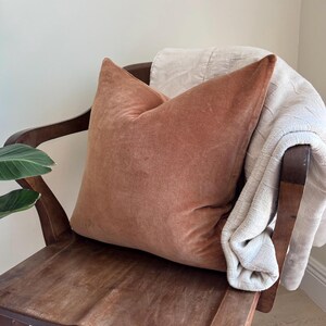 May include: A terracotta-colored velvet pillow rests on a dark wooden chair, with a cream-colored blanket draped over the back. The pillow has a soft, textured appearance, and the chair has a classic design.