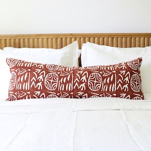Puede incluir: Un cojín lumbar rectangular largo de tela color terracota profundo con un patrón blanco repetitivo estampado a mano de motivos circulares y formas abstractas, colocado horizontalmente sobre una cama con ropa de cama blanca.