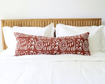 May include: A long rectangular lumbar pillow featuring a deep terracotta red fabric with a repeating white hand-printed pattern of circular motifs and abstract shapes, positioned horizontally across a bed with white bedding and a wooden headboard.