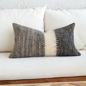 May include: A rectangular lumbar cushion featuring a textured, dark charcoal and grey woven fabric. The centre of the cushion is accented with a vertical, cream-coloured panel that displays a raised, stitched pattern with small, light-coloured fringe details.