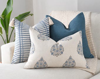 May include: Three decorative throw pillows arranged on a sofa. One pillow features a blue and white plaid pattern with corner tassels, one is solid dark blue, and the front pillow is off-white with a repeating blue floral print pattern.