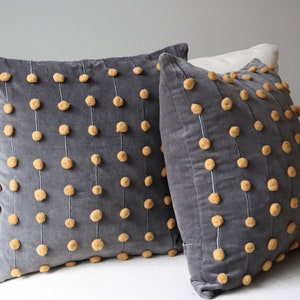 May include: Two grey velvet cushions with vertical rows of tan pom-poms. The pom-poms are evenly spaced and attached with white thread. The cushions are square and appear to be on a white surface.