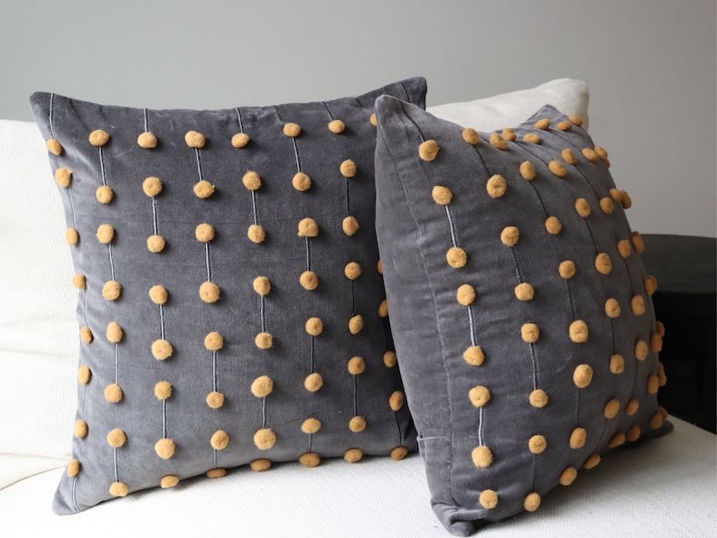 May include: Two grey velvet cushions with vertical rows of tan pom-poms. The pom-poms are evenly spaced and attached with white thread. The cushions are square and appear to be on a white surface.