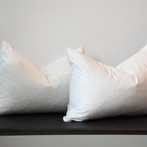 May include: Two white, fluffy pillows with a square shape are displayed on a dark wooden shelf. The pillows are plump and appear soft, with a tag visible on one. The background is a plain, light-colored wall.