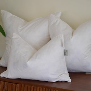 May include: Three fluffy white pillows are arranged on a wooden surface. The pillows have a soft, cloud-like appearance, with a tag on one pillow. The pillows are a neutral color and are likely for home decor.