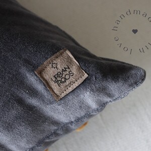 May include: Close-up of a charcoal grey cushion with a textured surface. A small, square, brown leather tag is sewn onto the cushion, with the words "URBAN TROOS" and a small graphic. The words "handmade with love" are in a circle around a heart.