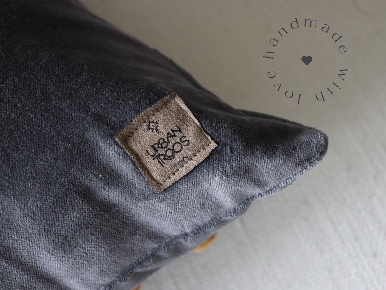 May include: Close-up of a charcoal grey cushion with a textured surface. A small, square, brown leather tag is sewn onto the cushion, with the words "URBAN TROOS" and a small graphic. The words "handmade with love" are in a circle around a heart.