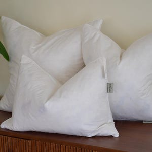 May include: Three fluffy white pillows are arranged on a wooden surface. The pillows have a soft, cloud-like appearance, with a tag on one pillow. The pillows are a neutral color and are likely for home decor.