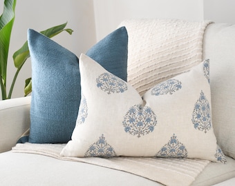 May include: Two decorative throw pillows on a cream-colored sofa. One pillow is a solid, textured blue. The other pillow is cream-colored with a blue floral pattern. A cream-colored blanket is draped over the sofa.