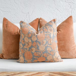 Puede incluir: Cuatro cojines decorativos. Dos son de color naranja quemado liso, y el cojín central tiene un estampado floral azul y naranja. Los cojines están colocados sobre una superficie blanca, con una pared blanca texturizada en el fondo.