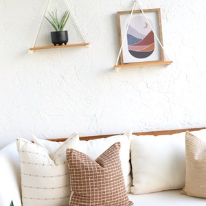 May include: Two wooden shelves with rope hangers, one with a potted succulent and the other with a framed abstract print of a landscape with a crescent moon. The shelves are hanging on a white textured wall above a white couch with brown patterned pillows.