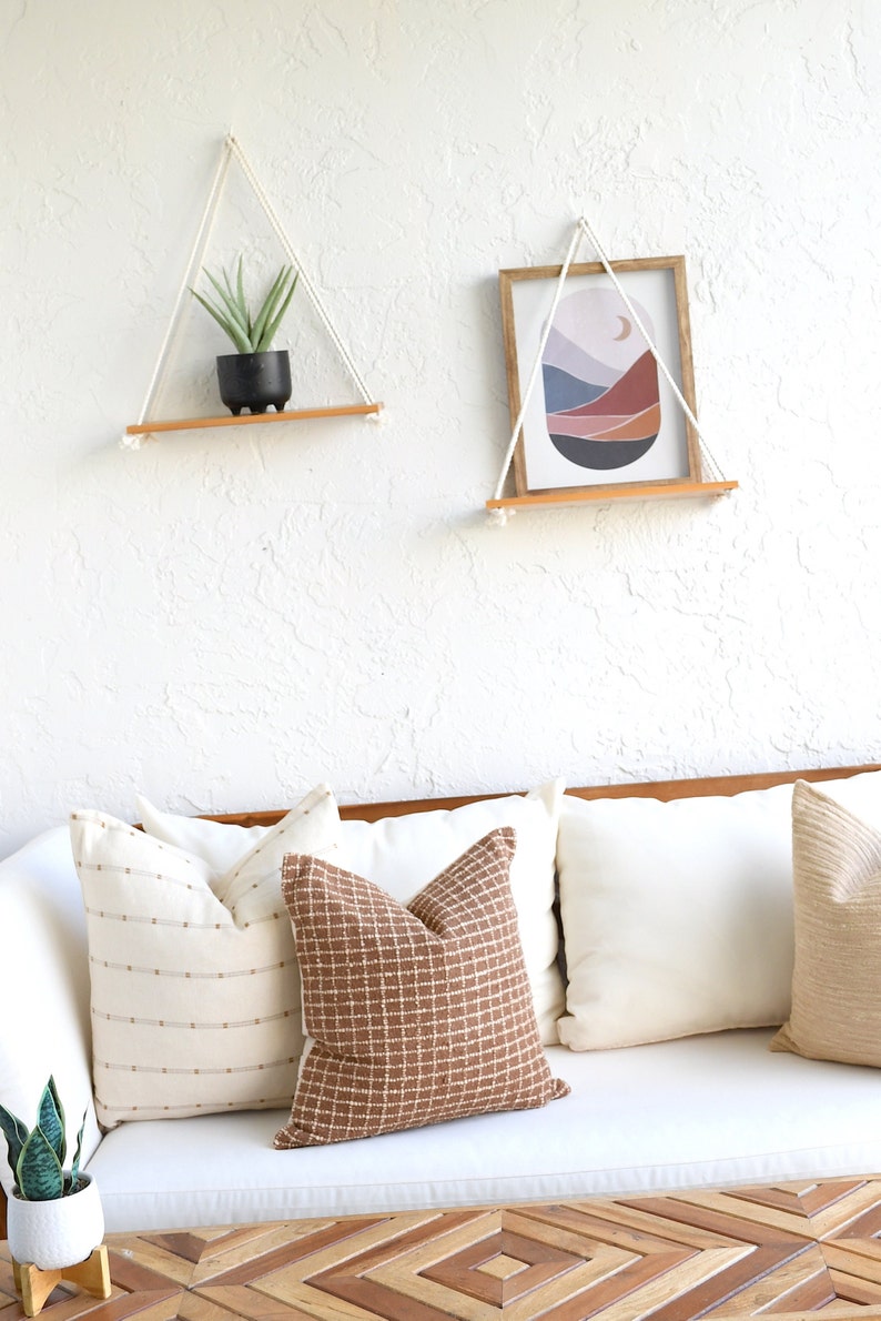 May include: Two wooden shelves with rope hangers, one with a potted succulent and the other with a framed abstract print of a landscape with a crescent moon. The shelves are hanging on a white textured wall above a white couch with brown patterned pillows.