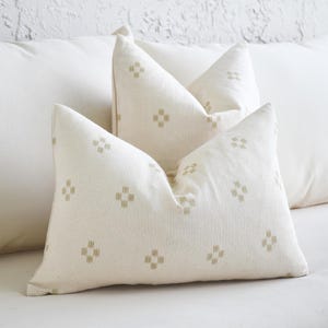 May include: Cream-colored throw pillows with a repeating geometric pattern in a muted green hue. The pillows are arranged on a white sofa, creating a neutral and inviting aesthetic. The pillows add a touch of texture and pattern to the space.